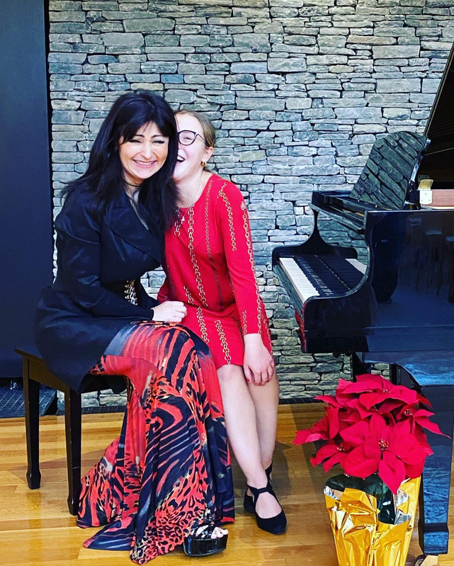 Two women seated by a piano, smiling in festive red and black outfits with poinsettias nearby