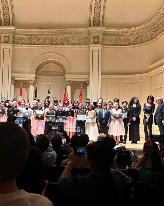 Choir on a grand stage receiving applause, with flags and ornate hall decor in the background.