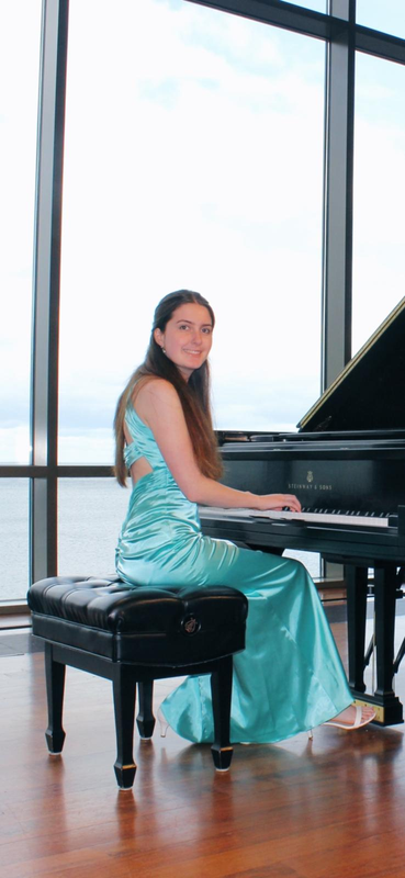 Woman in a turquoise dress seated at a grand piano in a bright room with floor-to-ceiling windows
