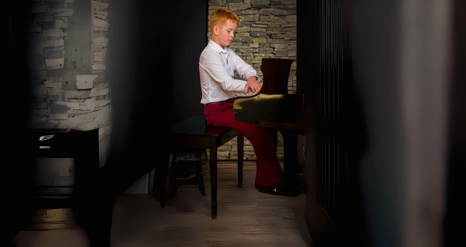 Person in a white shirt and red pants sits sideways on a stool in a dim hallway, arms crossed, facing left.