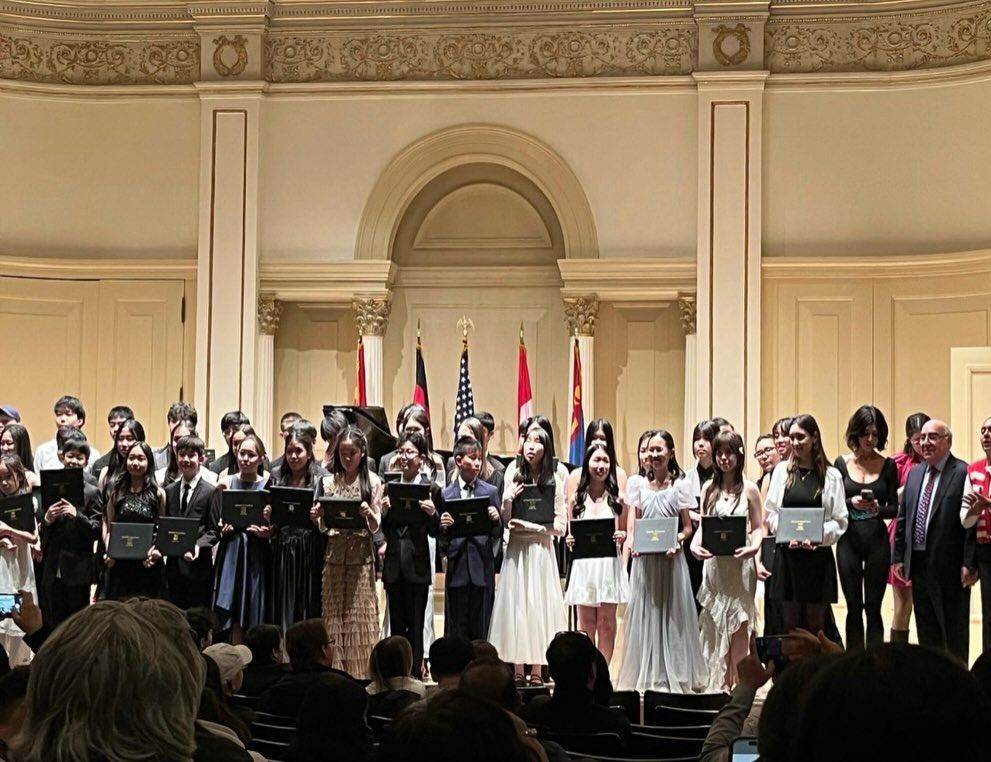 Choir members in formal attire stand on a stage in a grand hall, holding music folders.