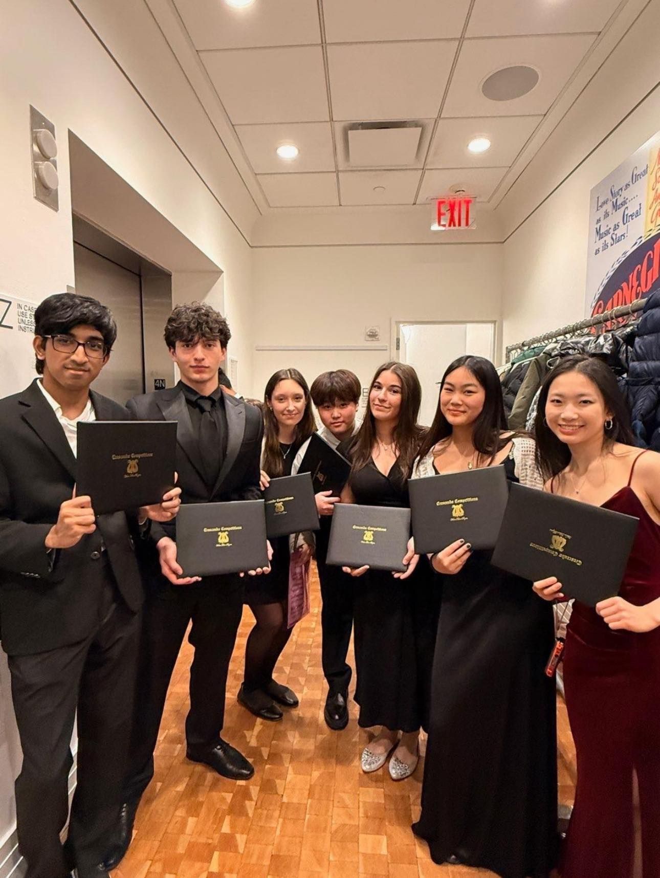 Group of six people in formal attire holding diplomas in a hallway, smiling at the camera.