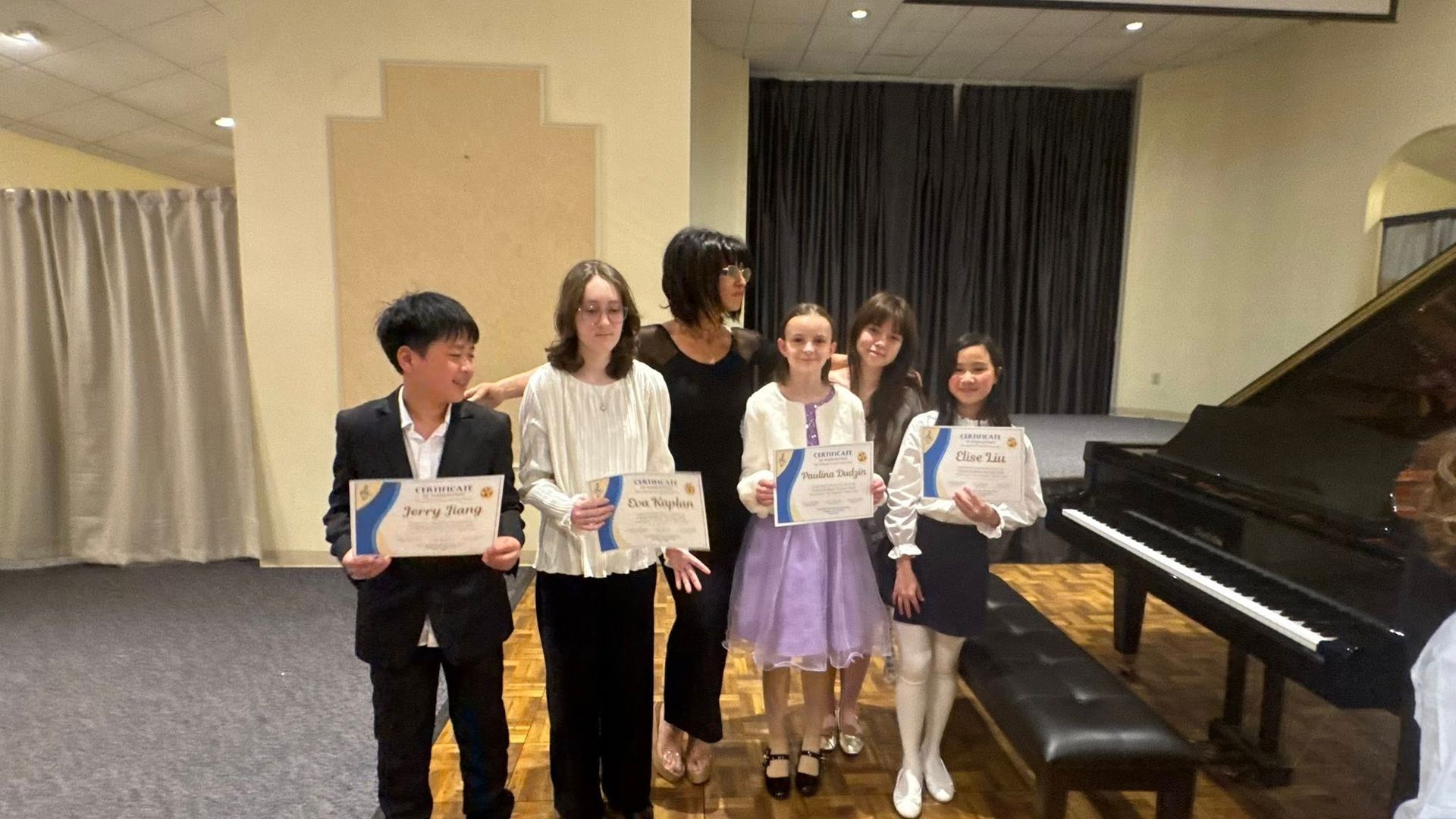 Five people holding certificates stand on a stage beside a grand piano.