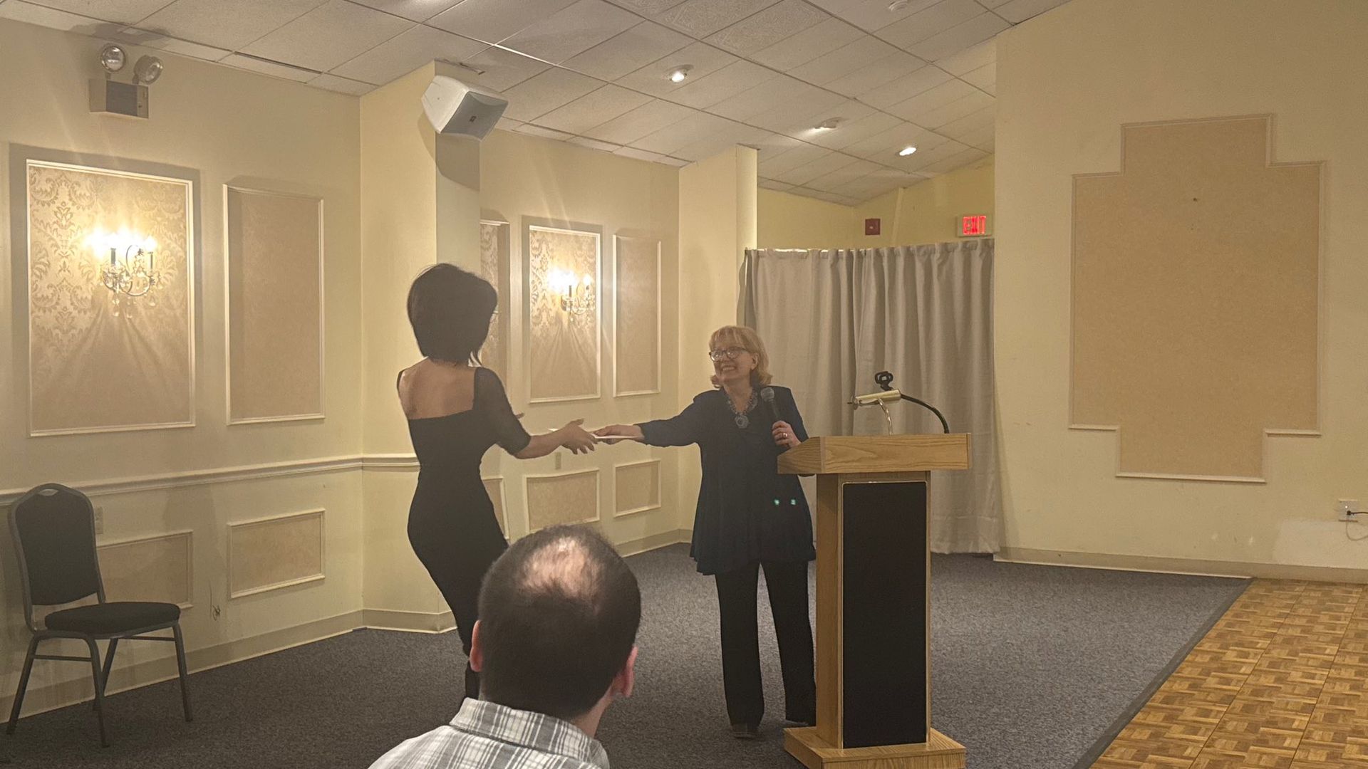 Two people speak at a podium in a banquet hall, with one standing beside it and an audience in front.