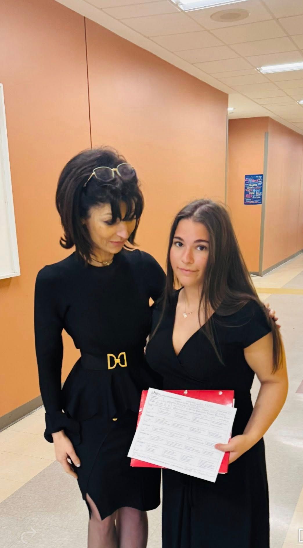 Two people standing in a hallway, one holding a notebook and pen, both wearing black outfits.