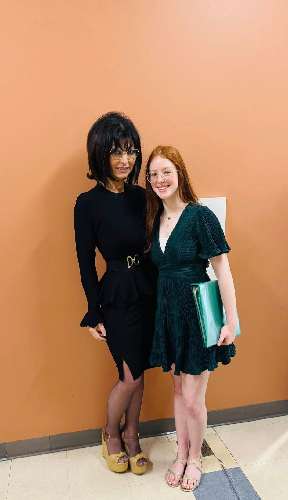 Two women posing together against an orange wall, one in a black dress and one in a green dress holding a folder