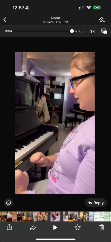 Person seated at a piano in a dim room, wearing glasses and a purple shirt, focused on playing keys.