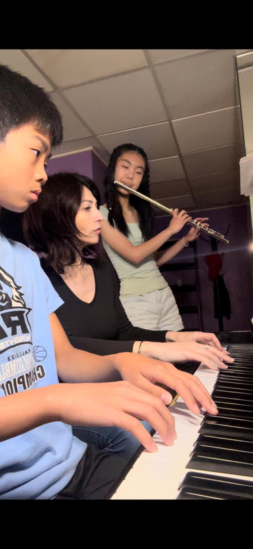 Three people playing instruments together in a dimly lit music room, including keyboard and flute.