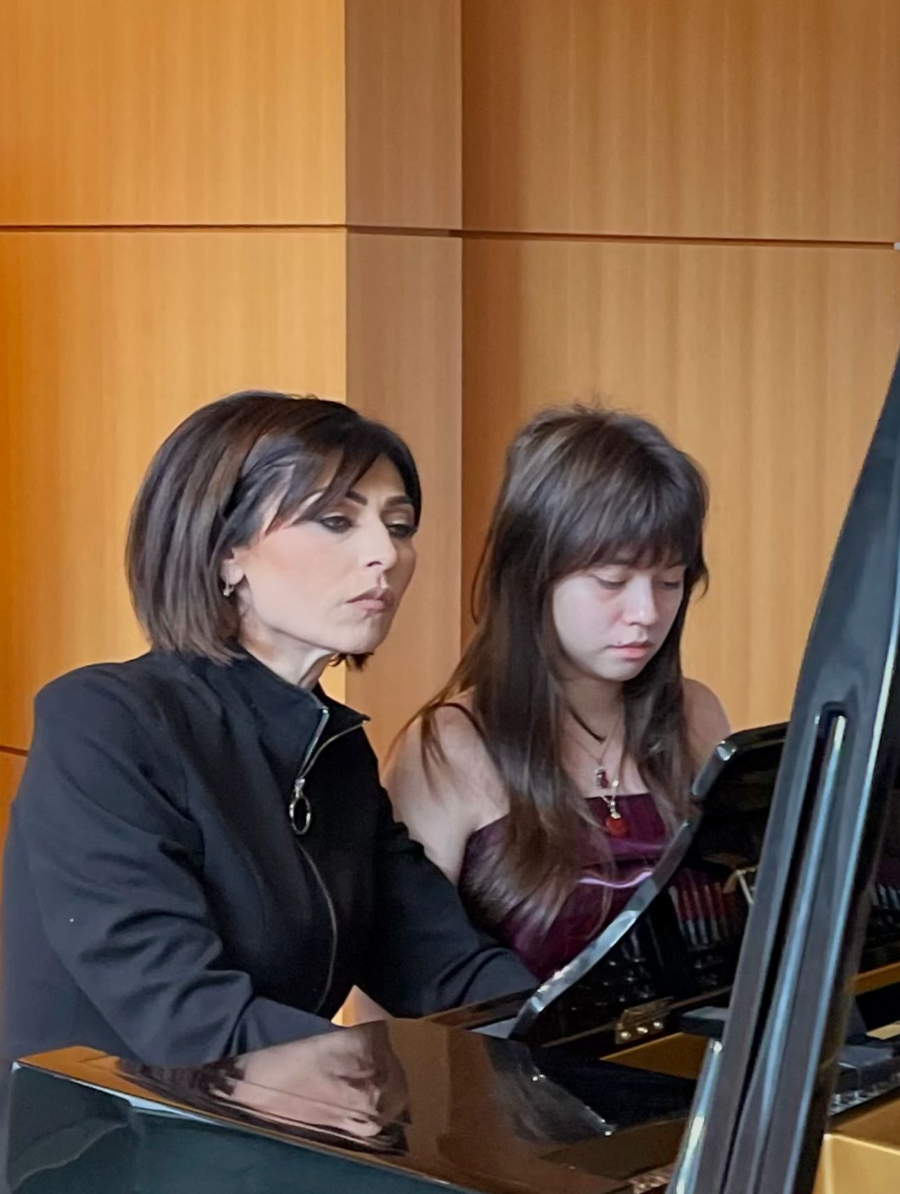 Two women seated at a piano, one singing or speaking while the other looks down at sheet music.