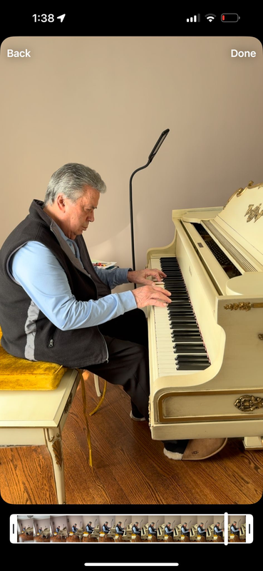 Man playing a cream-colored piano in a small room, viewed from behind, with a microphone nearby