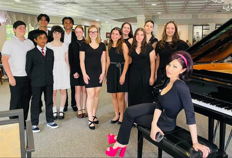 Group of people posing beside a grand piano in a bright music room, with one woman seated on the bench.