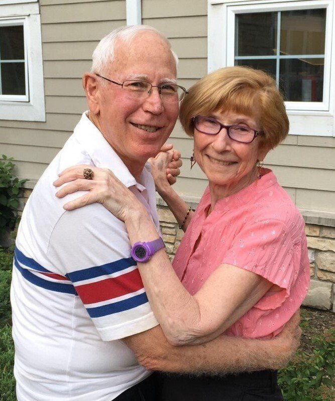 An elderly couple dances outdoors, smiling. The man wears glasses and a striped polo shirt. The woman has short red hair and a pink shirt.