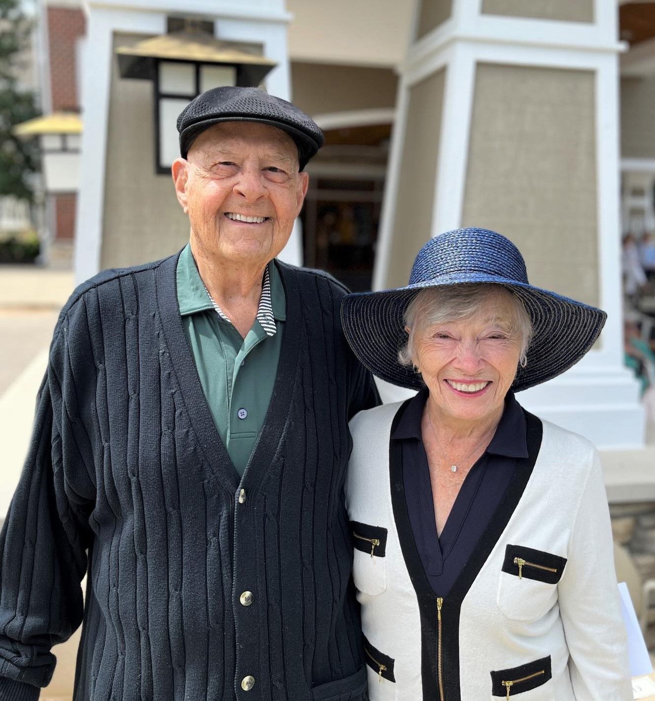 An elderly couple smiling, posing outdoors. Man wears a cap and cardigan, woman a wide-brimmed hat and jacket.