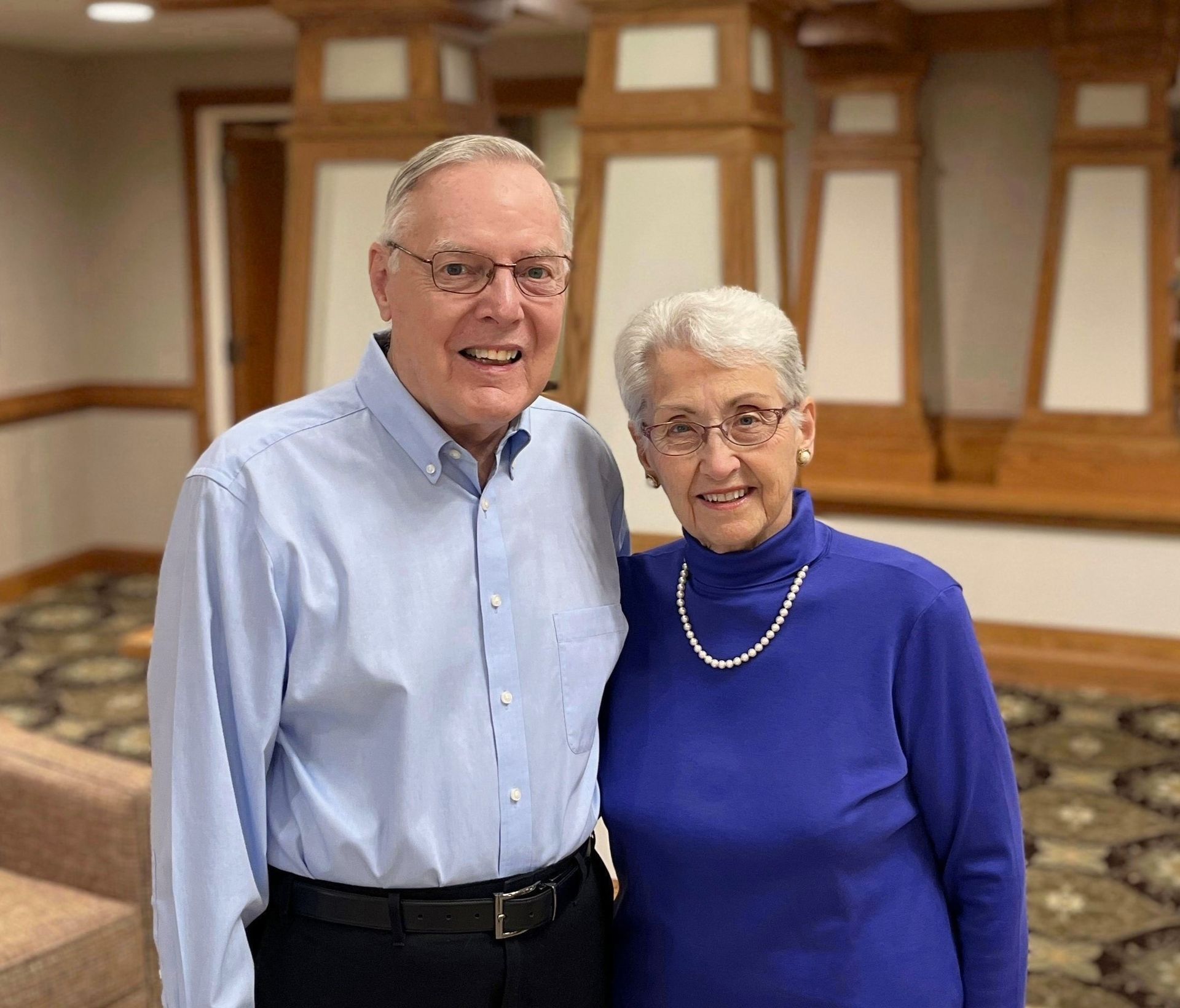 An older man and woman smile together, posing indoors. The man wears glasses and a blue shirt. The woman wears a blue turtleneck and a pearl necklace.