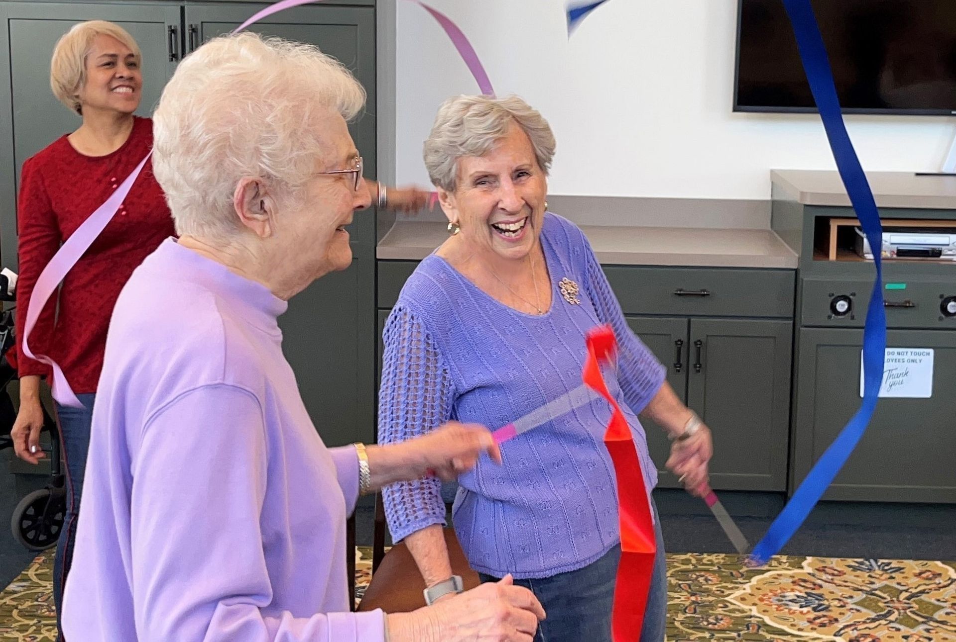Three older women smiling while cutting ribbons at an indoor event. Two women are cutting ribbons with scissors, another watches from the background.