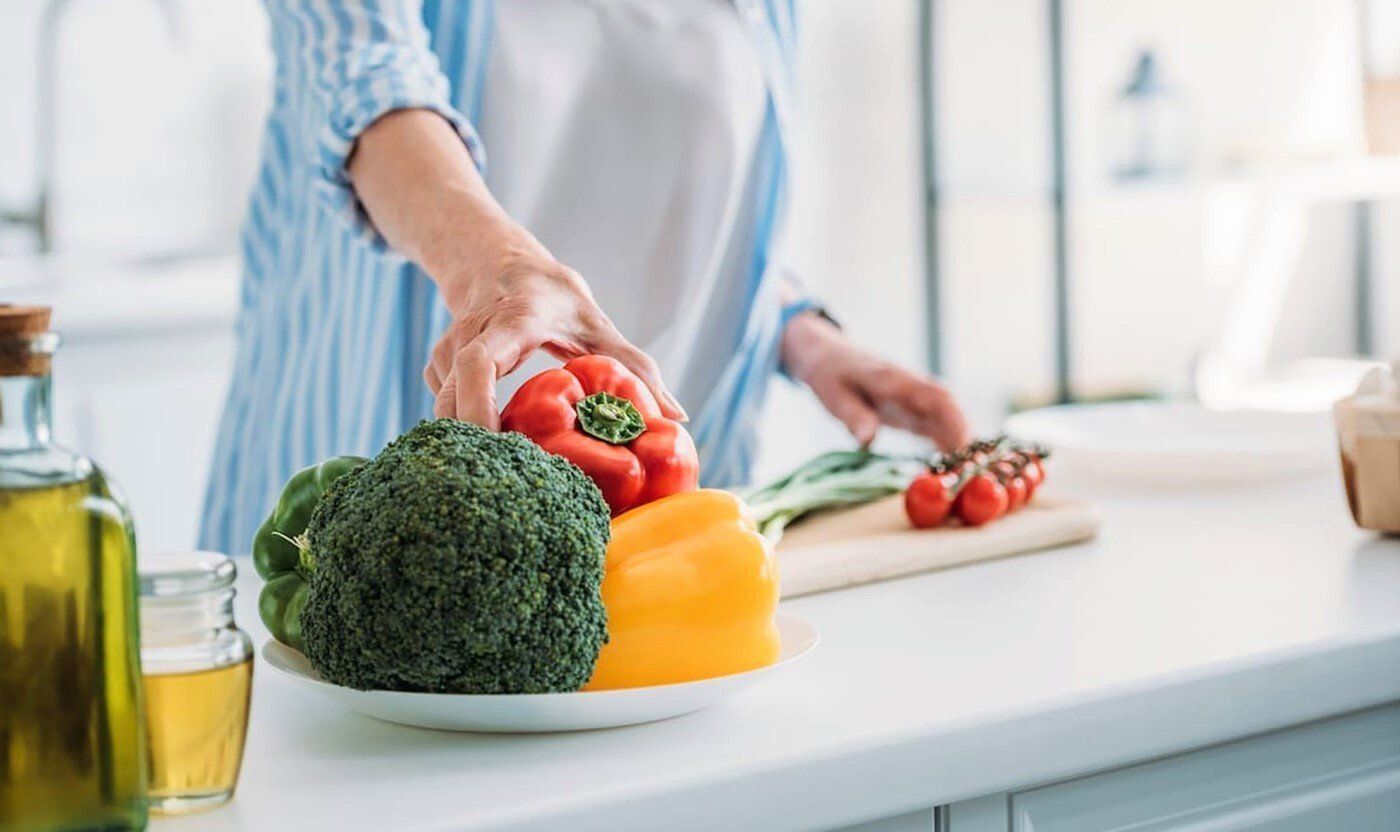 Person preparing vegetables in a kitchen. A plate of broccoli and bell peppers sits on the counter, with a cutting board and olive oil nearby.