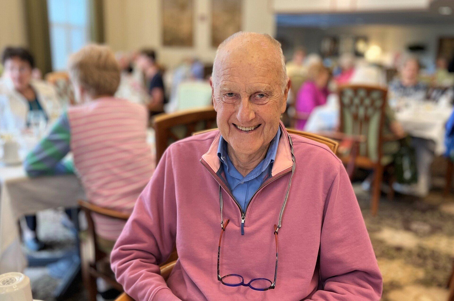 An elderly man with a bald head and pink sweater smiles at the camera in a dining room with other people.