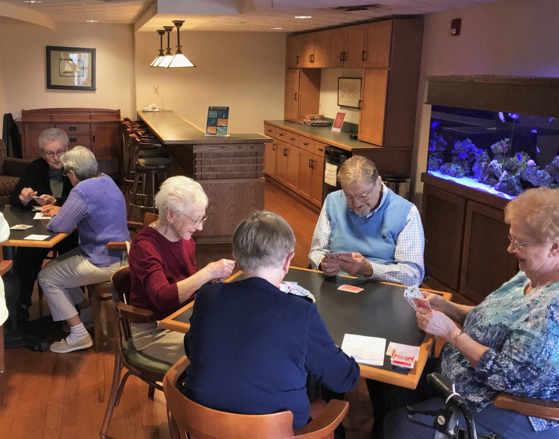 Group of seniors playing cards at tables in a community room. Some are smiling, and a large aquarium sits in the background.