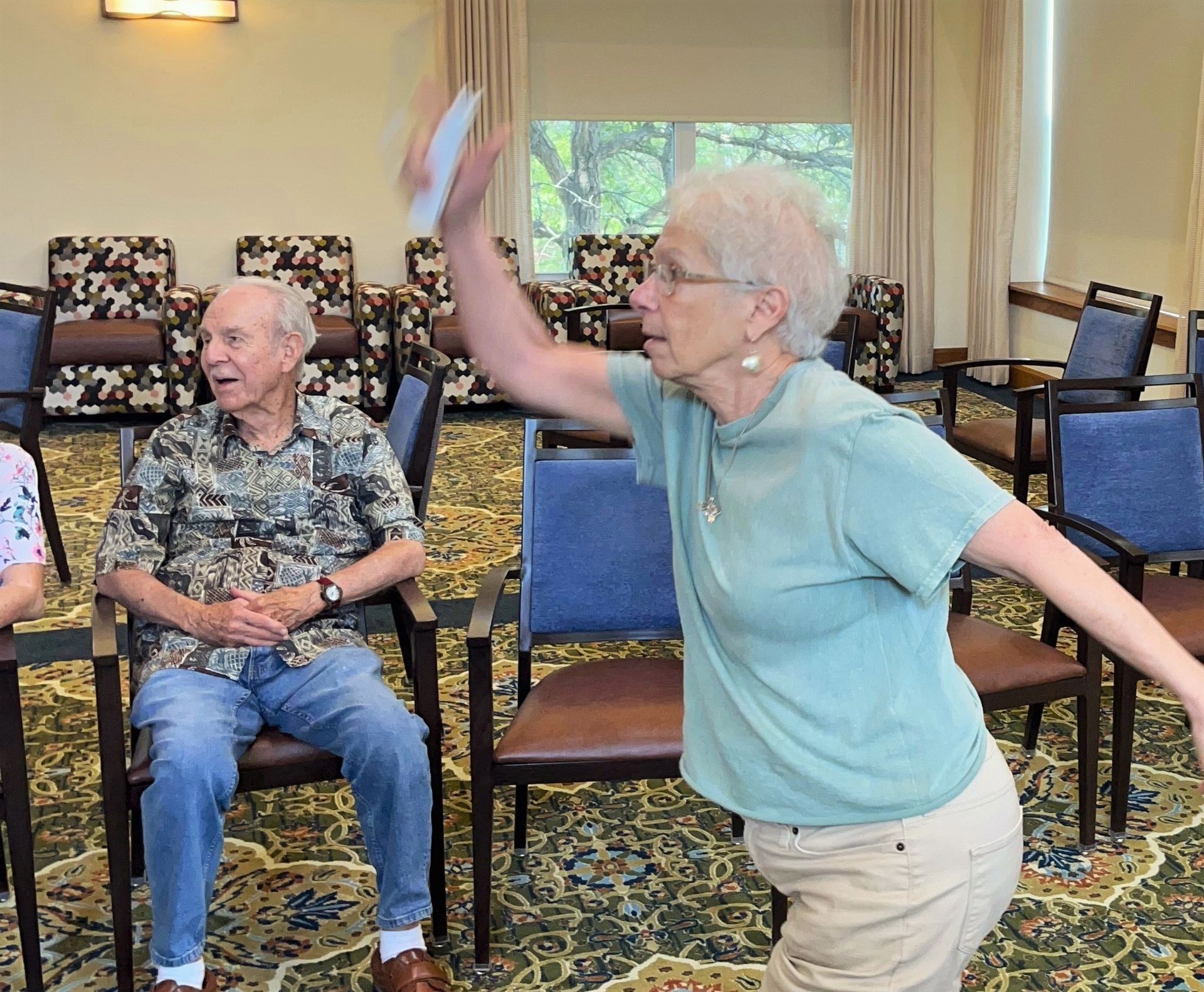 A woman in a light green shirt throws something in the air, while an older man watches from a chair indoors.