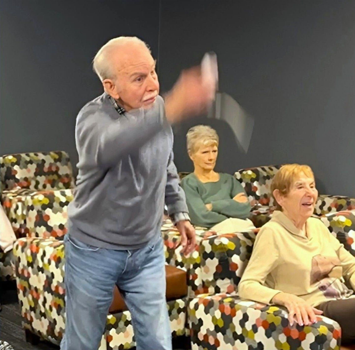 Elderly man gesturing excitedly, standing near patterned armchairs. Two other seniors sit, observing, one with arms crossed.