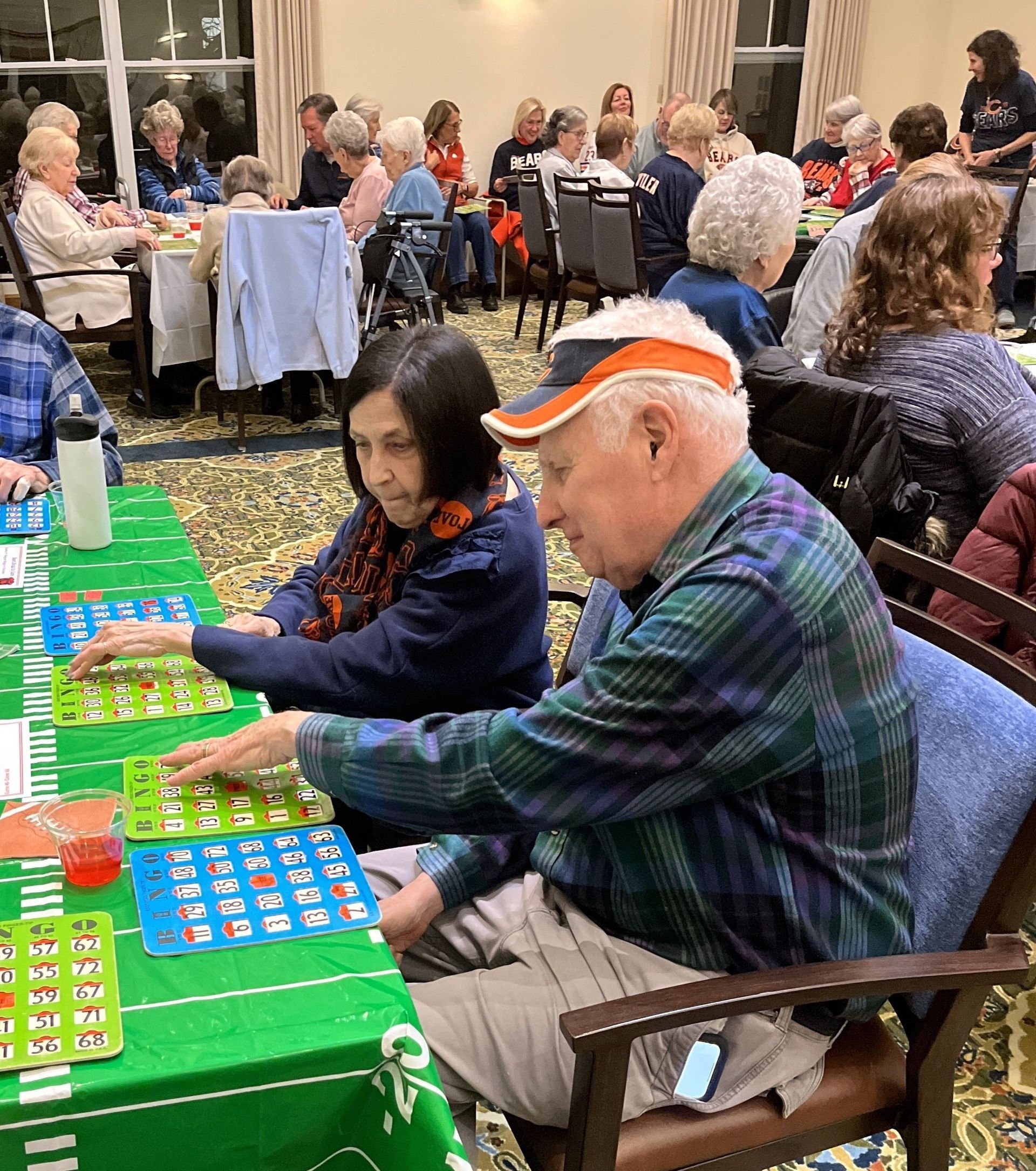 People playing bingo at a table in a community center. A man in a cap and a woman in a scarf mark their cards, surrounded by other players.