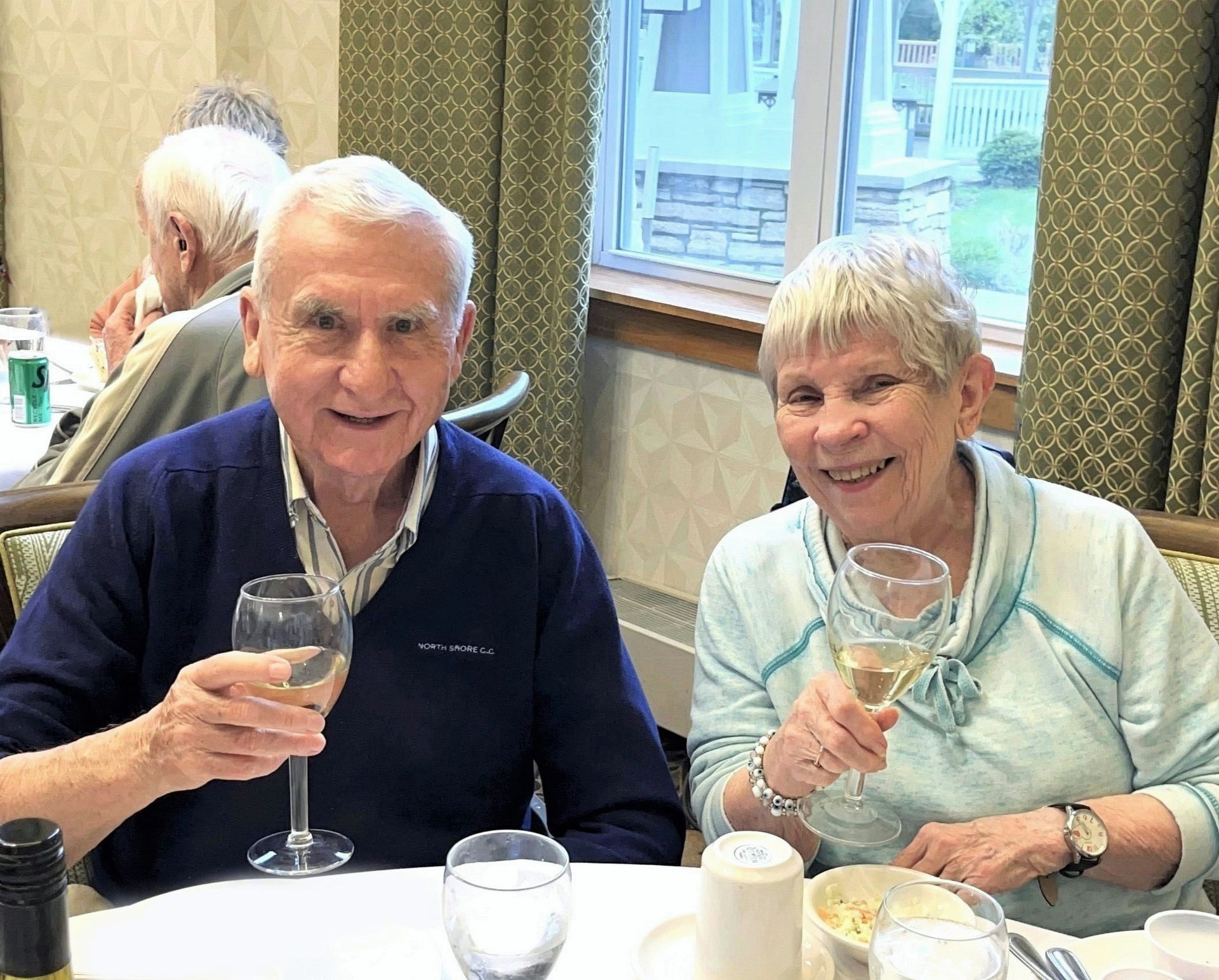 Elderly couple at a table, toasting with wine glasses. The man wears a blue sweater, the woman a blue sweater. They are smiling.