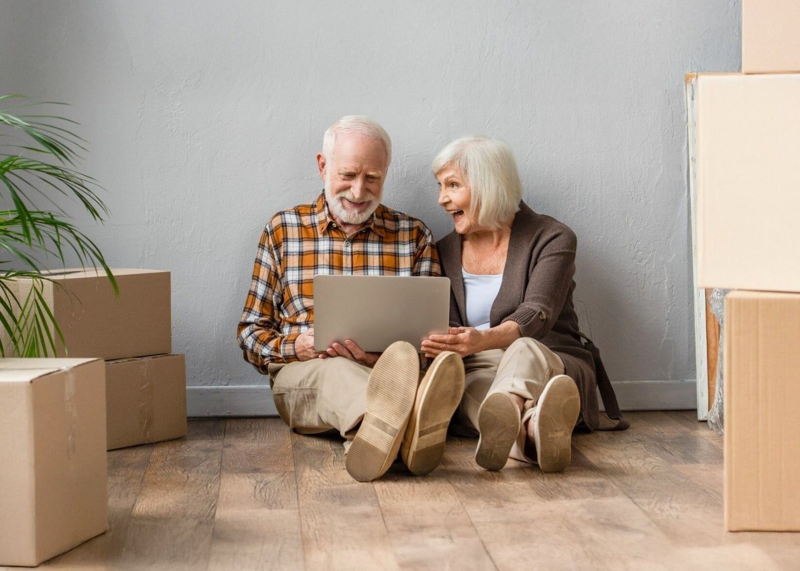 Elderly couple sitting on the floor, looking at a laptop, surrounded by moving boxes. They appear happy, smiling.
