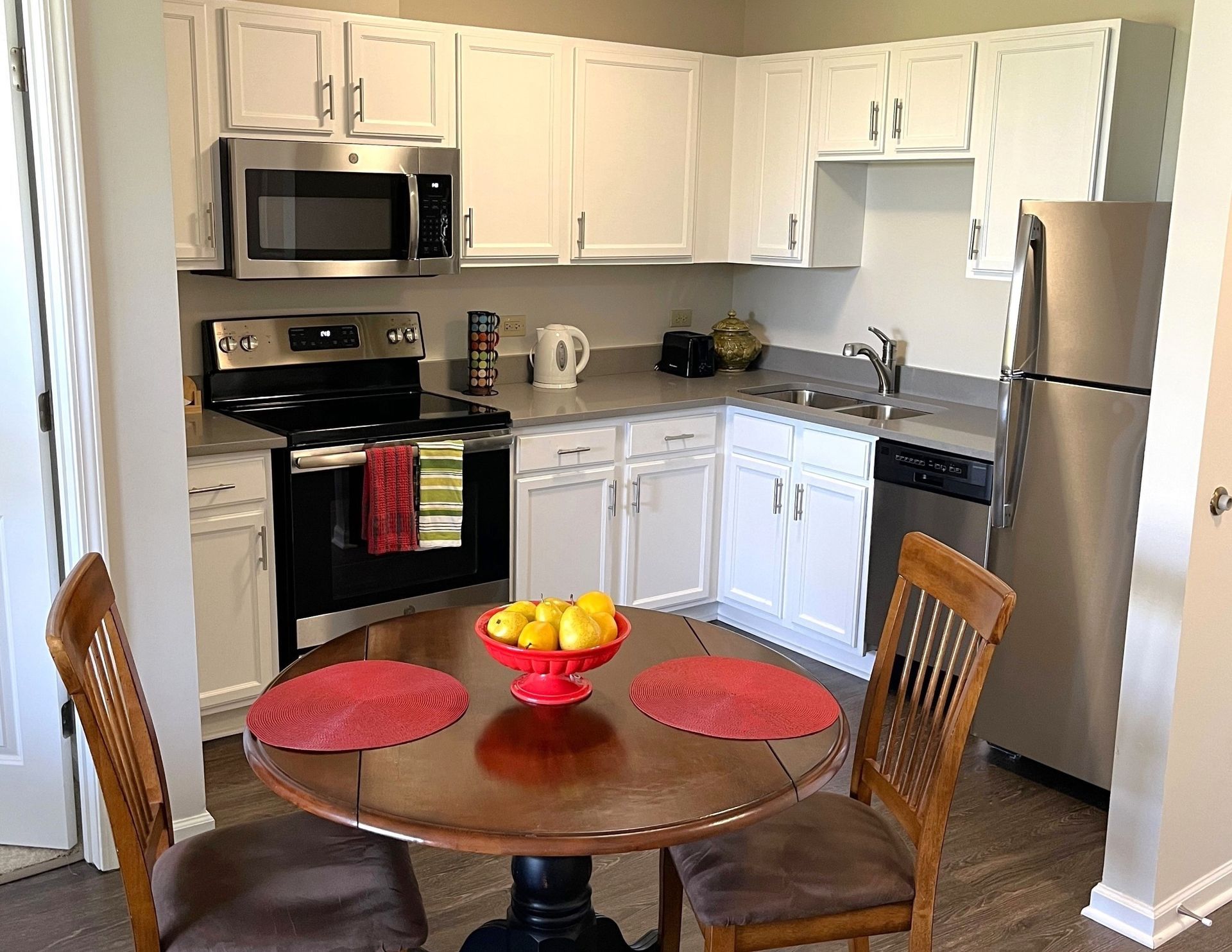 Kitchen with white cabinets, stainless steel appliances, a round wooden table set for two with fruit centerpiece.