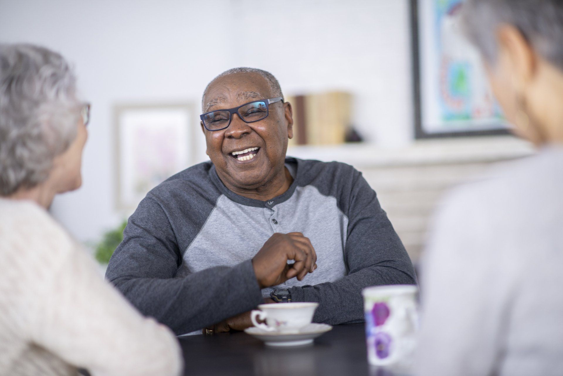 A smiling Black man in glasses chats with two people at a table. He wears a gray shirt, holding a cup.