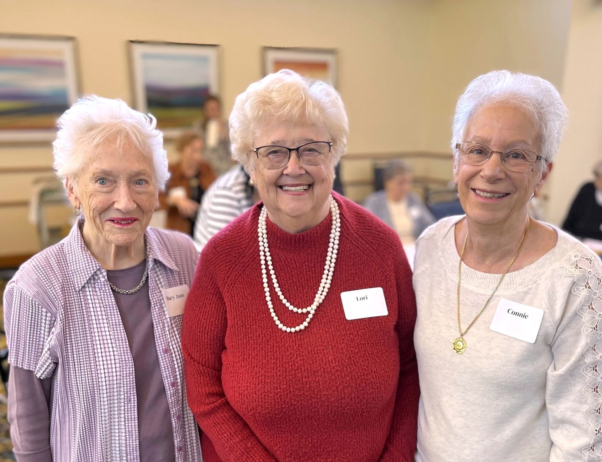 Three smiling older women with white hair stand together indoors. They wear sweaters and jewelry, posing for a photo.