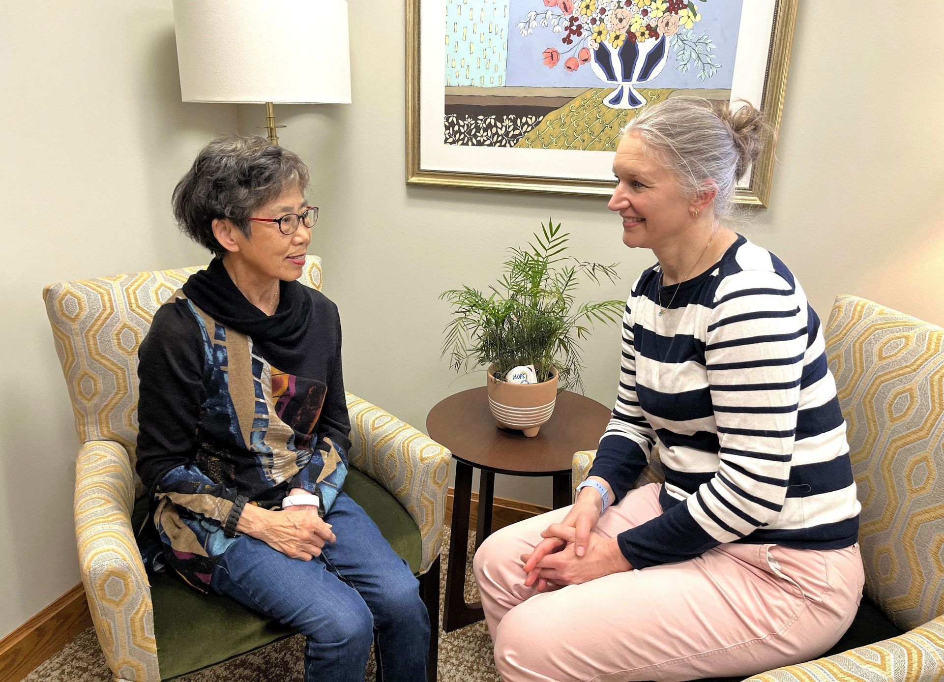 Two women seated in armchairs, facing each other. One wears a striped shirt and pink pants, smiling. The other wears a patterned top and jeans. They are in a room with artwork and a small table.