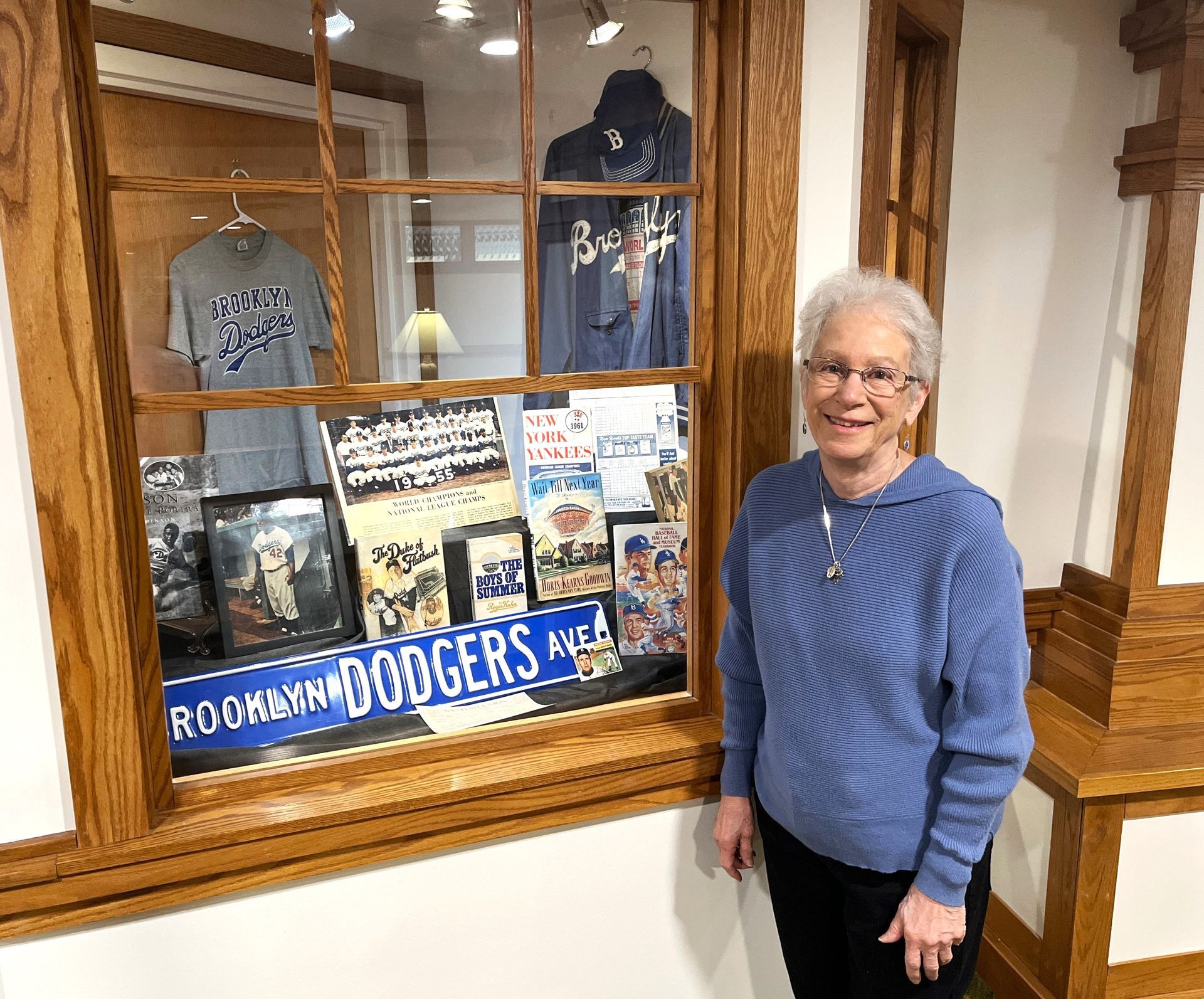 Woman stands by a display case with Brooklyn Dodgers memorabilia, including shirts, signs, and photos. She wears a blue sweater and smiles.