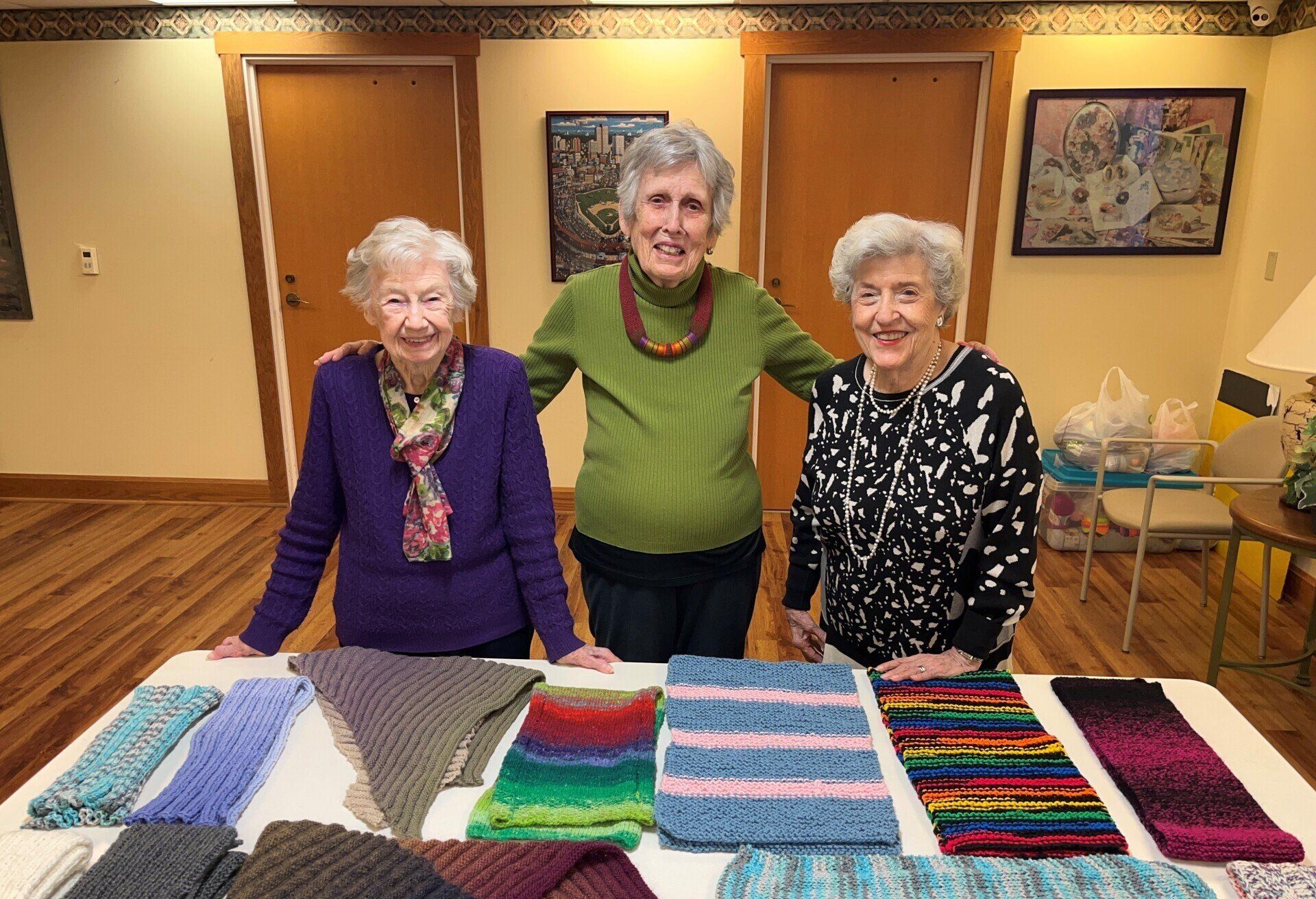 Three older women stand behind a table displaying colorful knitted scarves. They are smiling in a room with art and wooden doors.