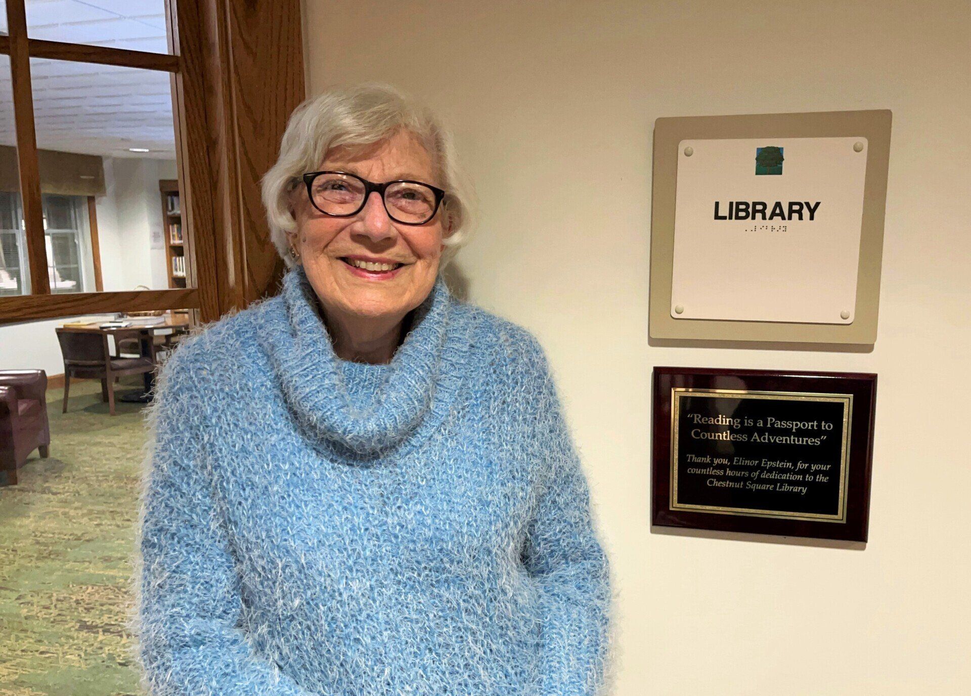 A smiling older woman with glasses, wearing a blue sweater, stands next to a