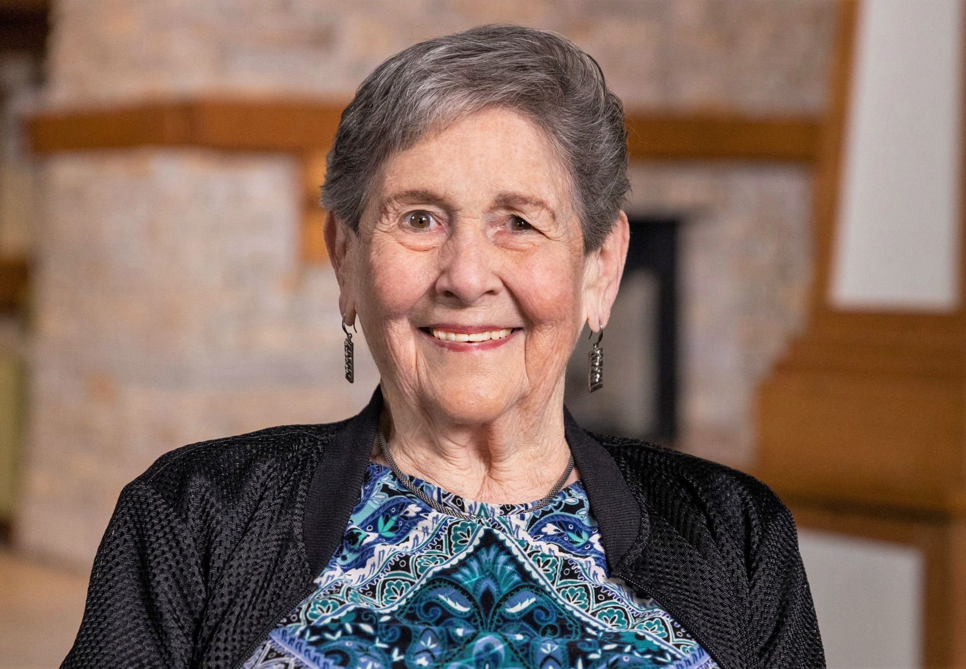 Smiling elderly woman with short gray hair, wearing a patterned top and black cardigan, indoors.