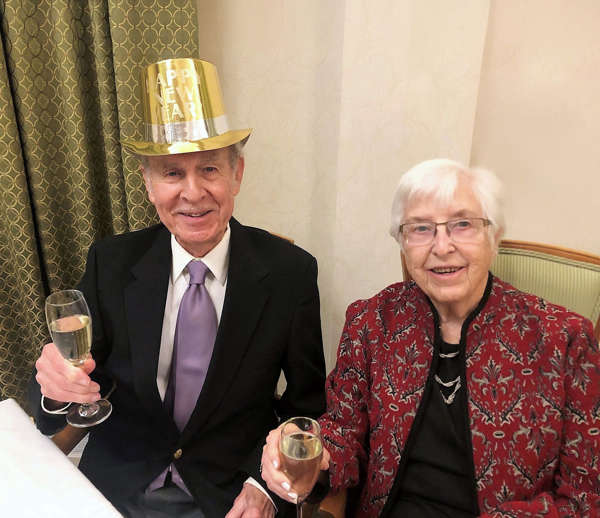 An older couple celebrating. The man wears a gold top hat and holds a champagne glass. The woman wears a red patterned jacket and also holds a champagne glass.