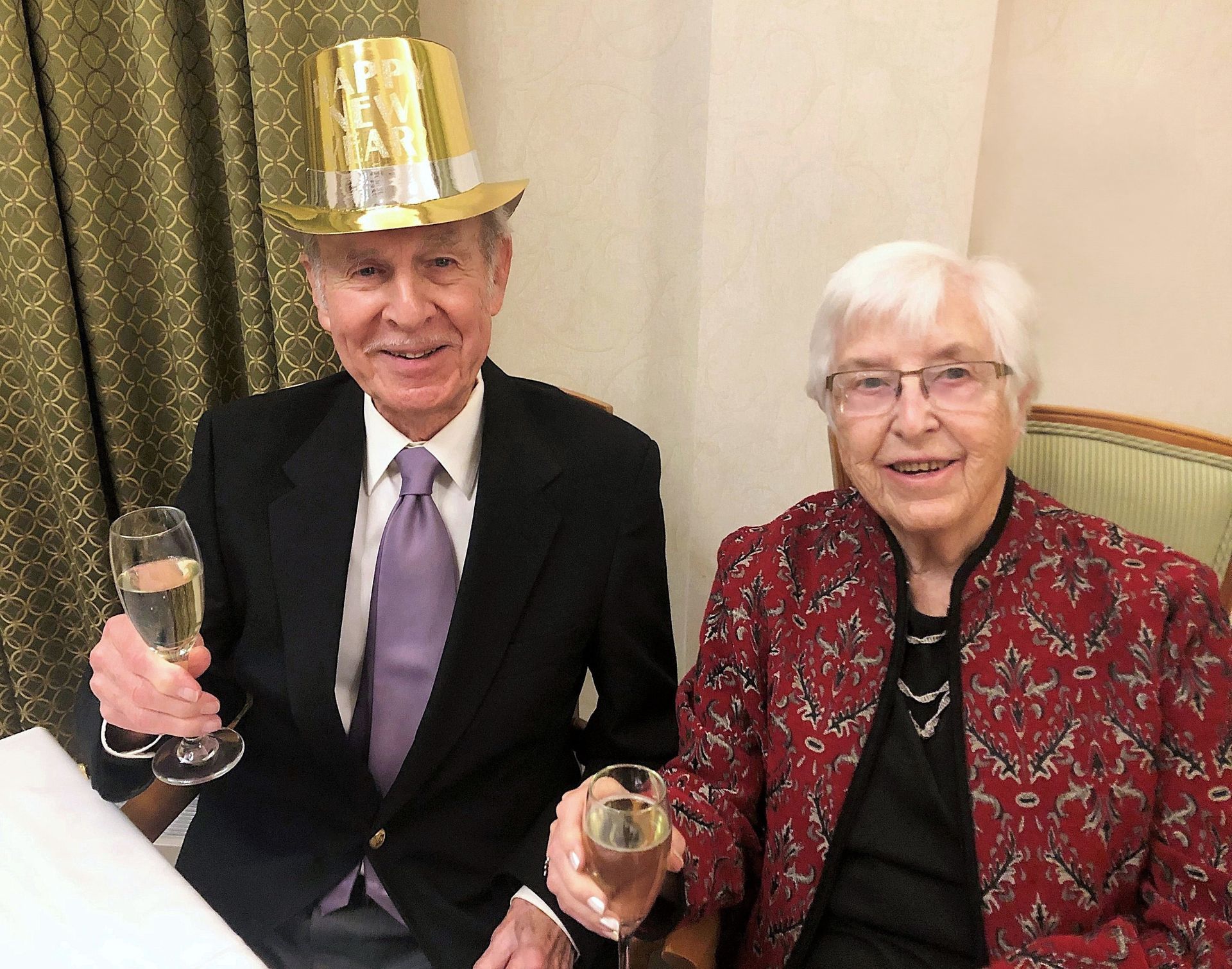 An elderly couple toasting with champagne. The man wears a top hat and suit, while the woman wears a red patterned jacket and glasses, all indoors.