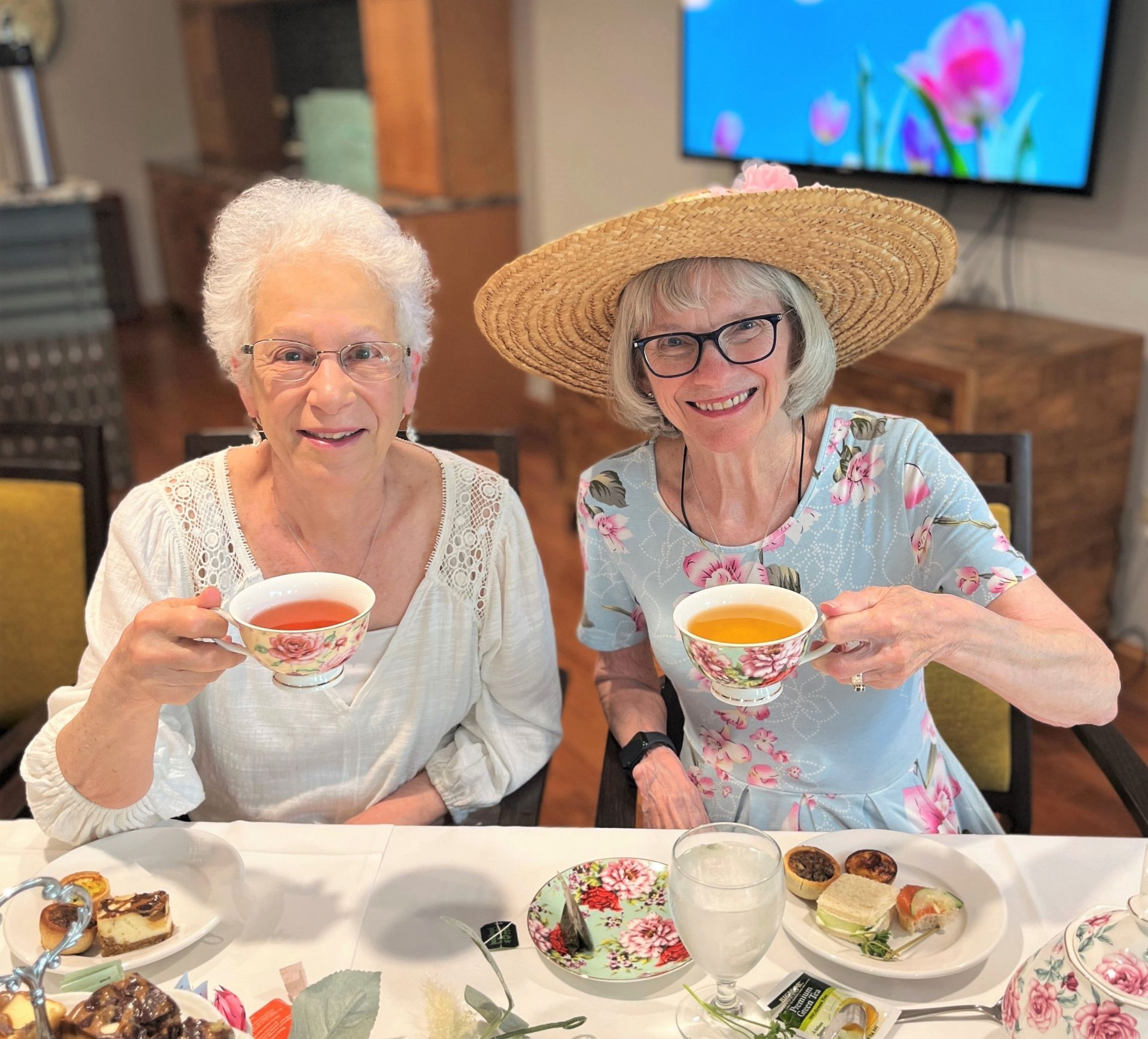 Two older women smile, enjoying tea at a table set with snacks and a floral tea set. One wears a straw hat, the other has white hair.