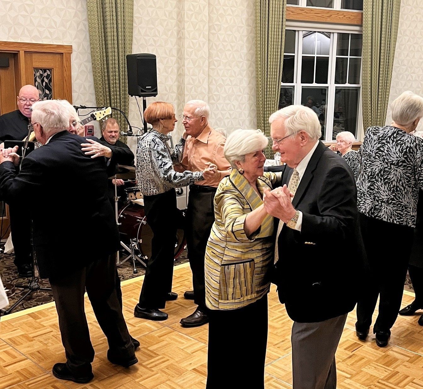 A group of senior citizens dances on a wooden floor to music at a social event. A band plays in the background.