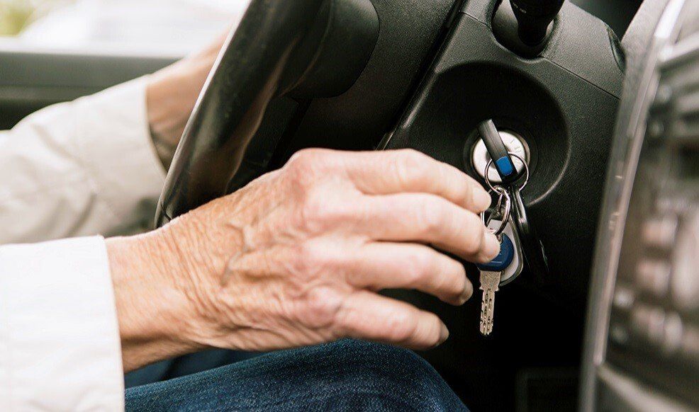 Close-up of older person's hands inserting car keys into the ignition, with the steering wheel in view.