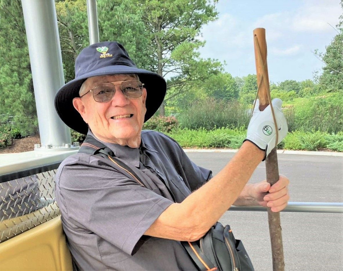 An elderly man in a hat and sunglasses smiles, holding a walking stick and wearing a golf glove; he's seated on a cart outdoors.