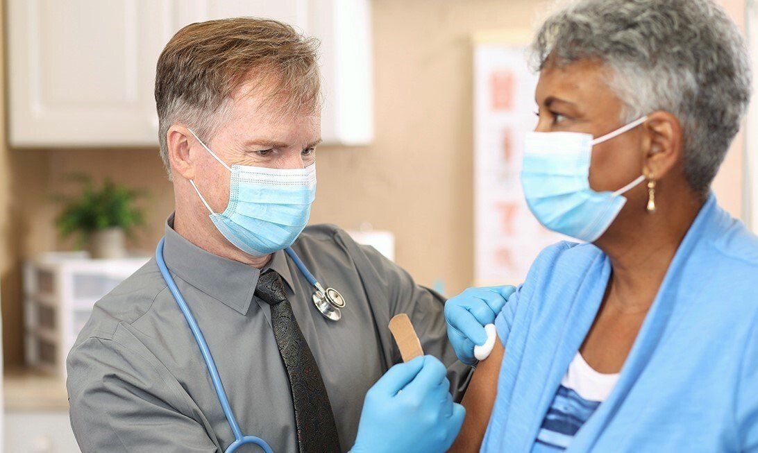 Doctor in mask and gloves giving an older woman in a mask a shot in the arm in a medical office.