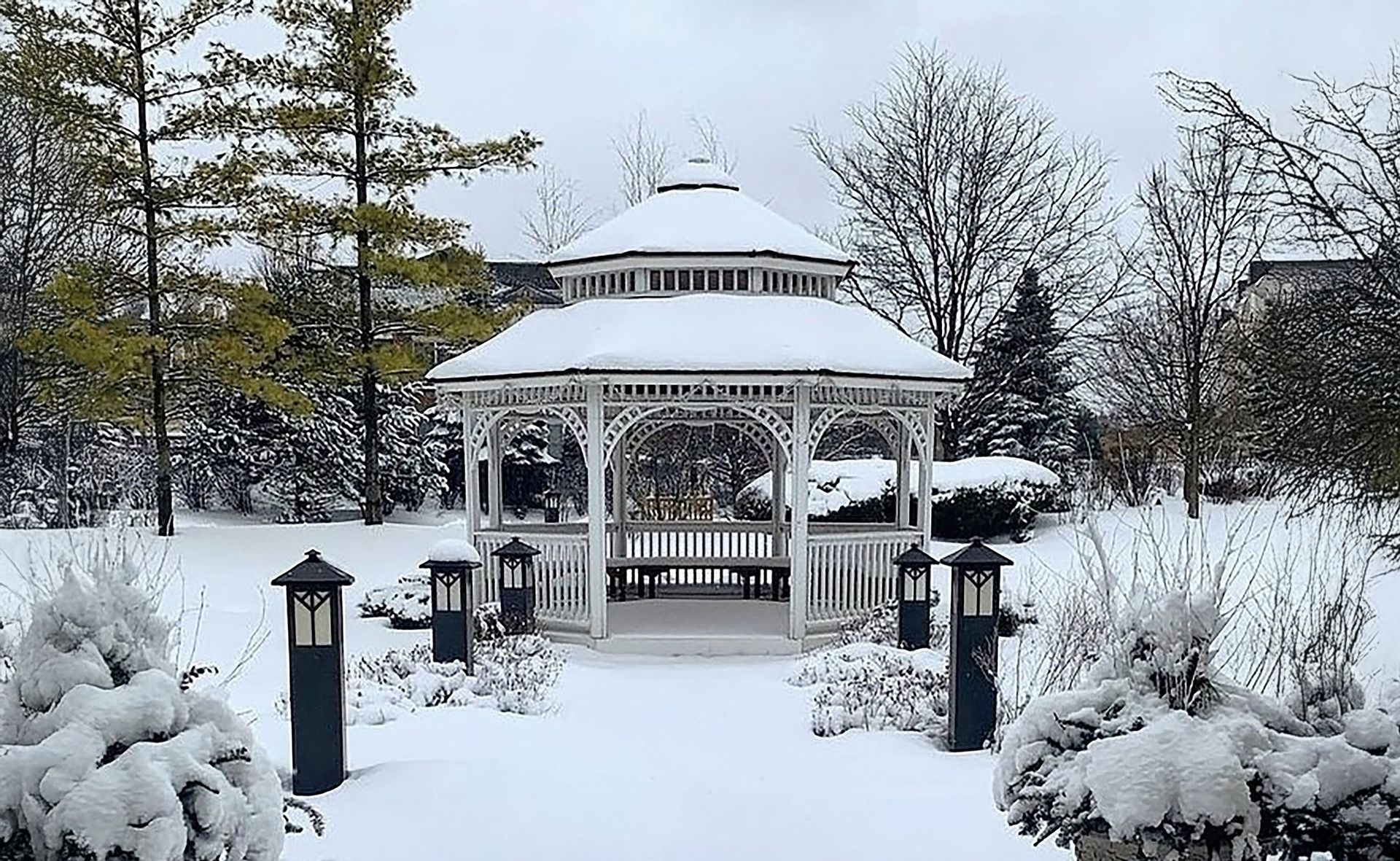 A snow-covered gazebo in a winter garden. White structure with path lined by lanterns and snow-dusted bushes and trees.