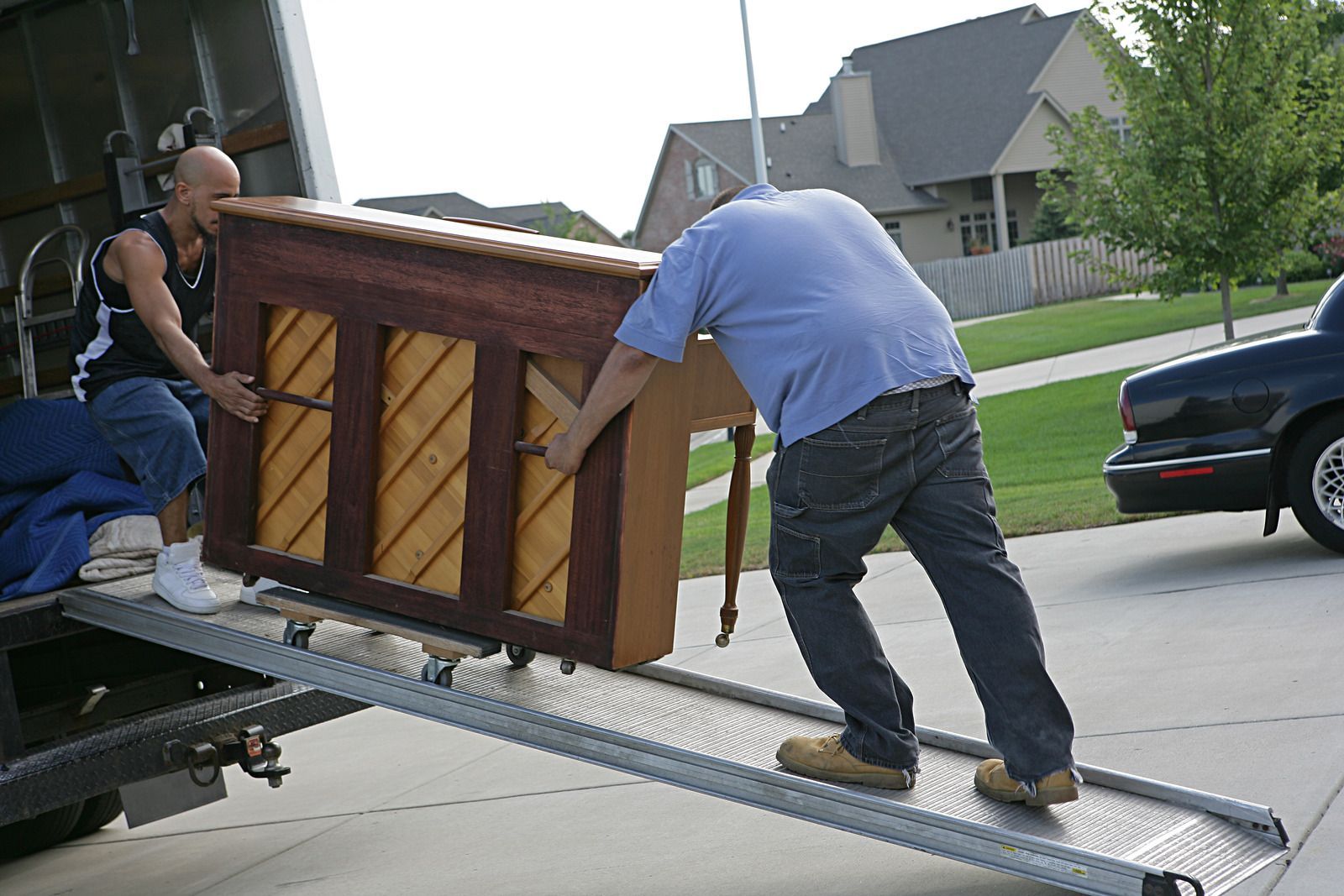 Two men are carrying a piano on a ramp from a truck