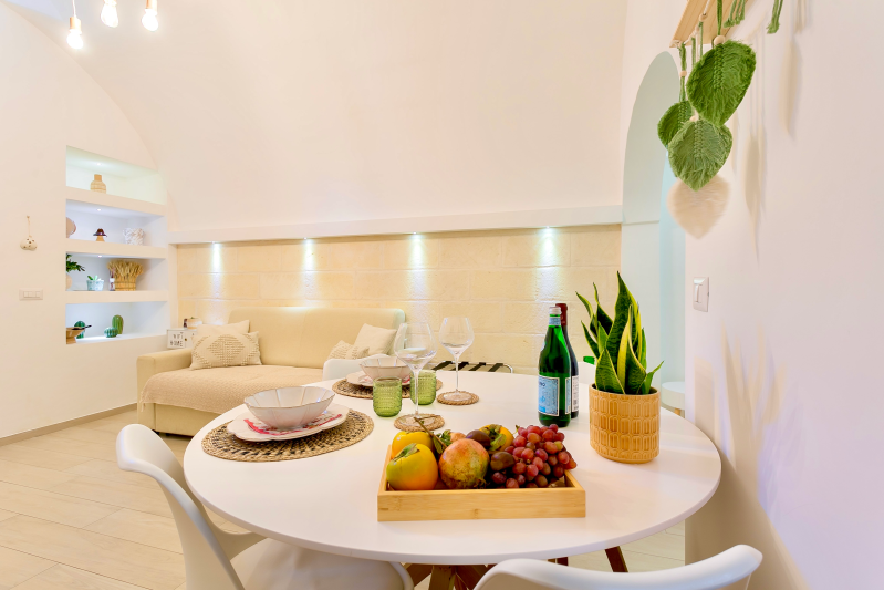 Interno di una sala da pranzo con pareti bianche, un tavolo rotondo apparecchiato per mangiare, frutta e un divano sullo sfondo.