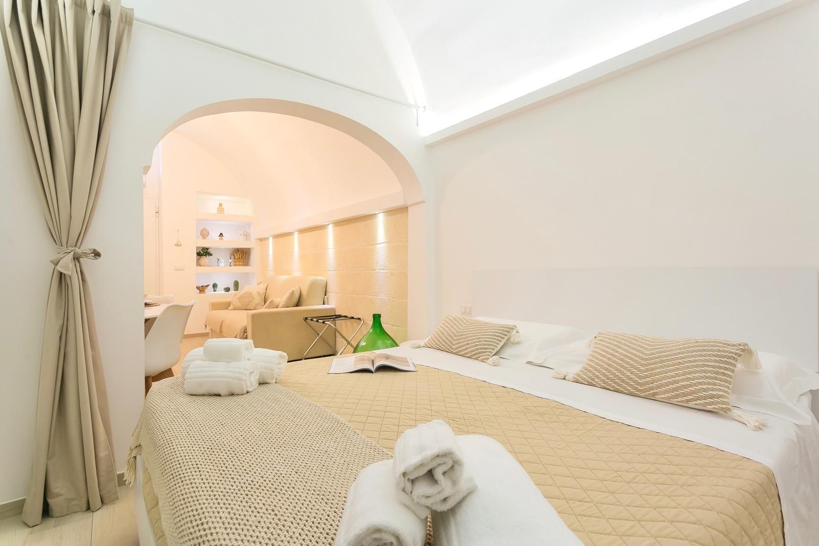 Interno della camera da letto con arco che conduce alla zona giorno, pareti bianche, biancheria da letto color crema, asciugamani e un libro.