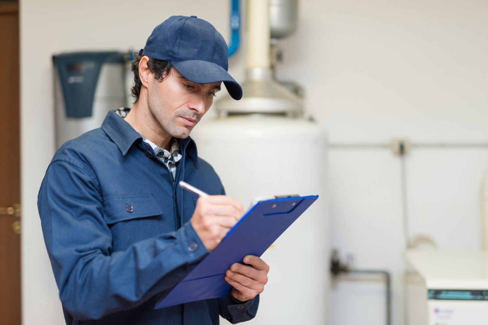 Technician in blue coveralls and cap inspects equipment, writing on a clipboard in a utility room.