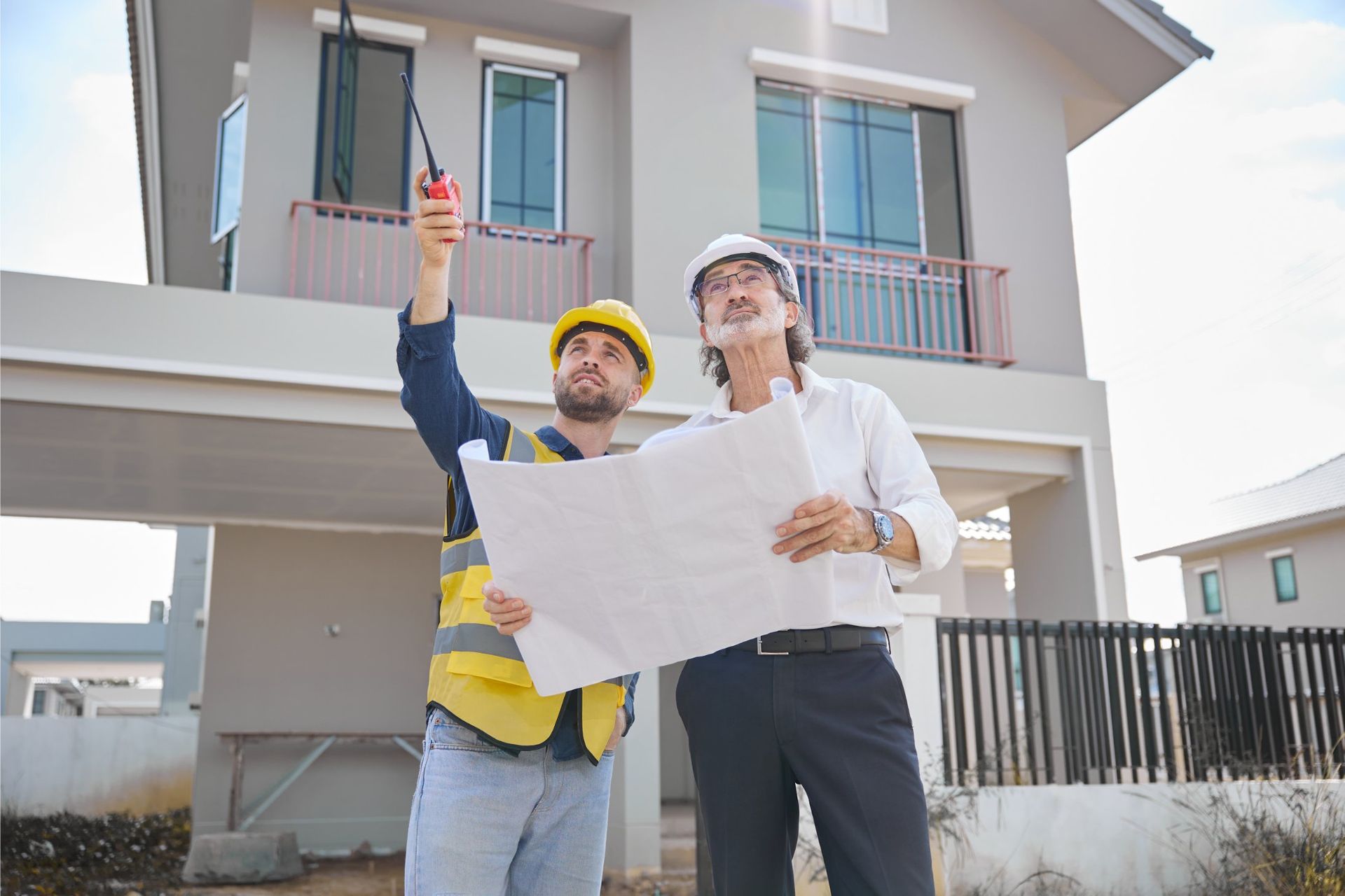 Two men at a construction site reviewing blueprints. One points up, holding a radio; the other wears a hard hat.