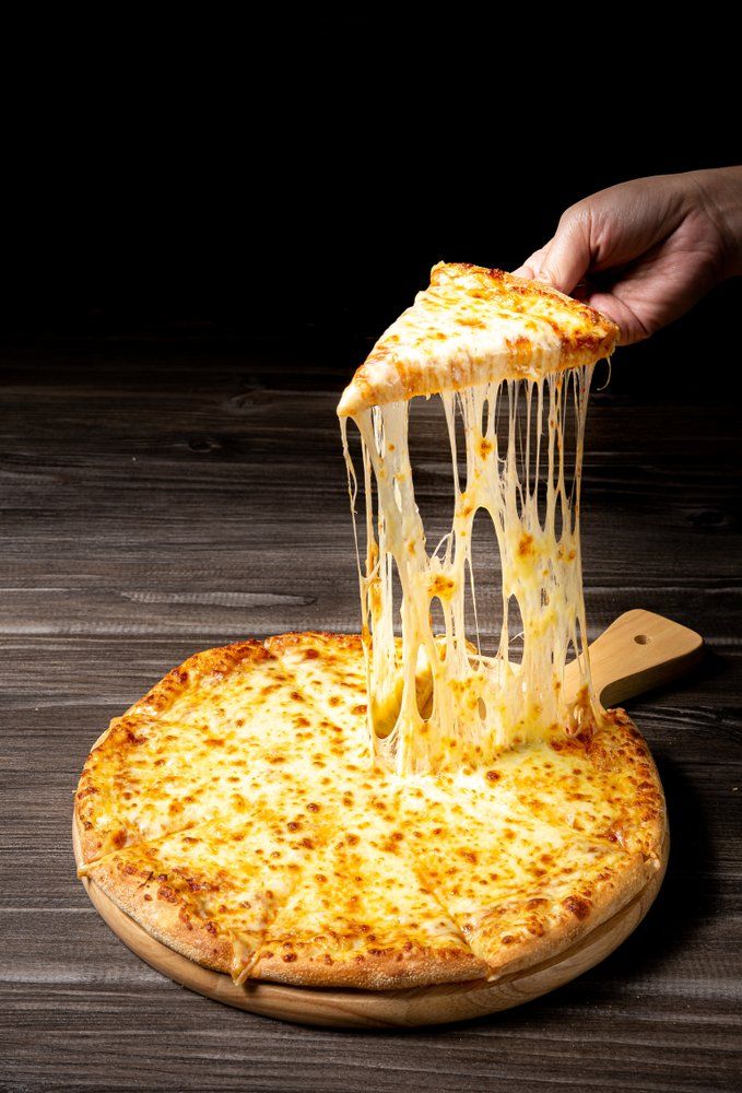 Une main soulève une part de pizza bien garnie de fromage, le fromage recouvrant toute la pizza posée sur une planche en bois.
