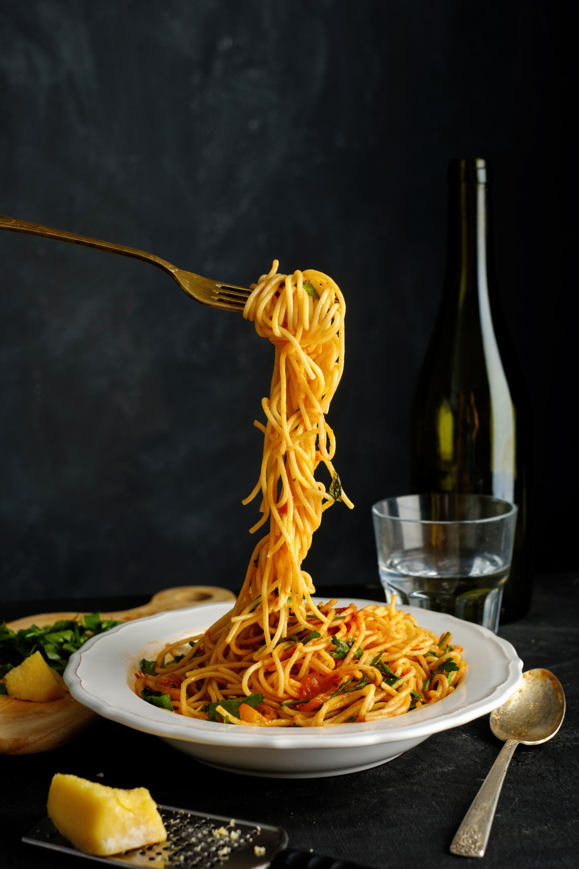 Plat de spaghettis avec une fourchette retenant un spaghetti. Bouteille, verre, fromage et cuillère sur fond sombre.